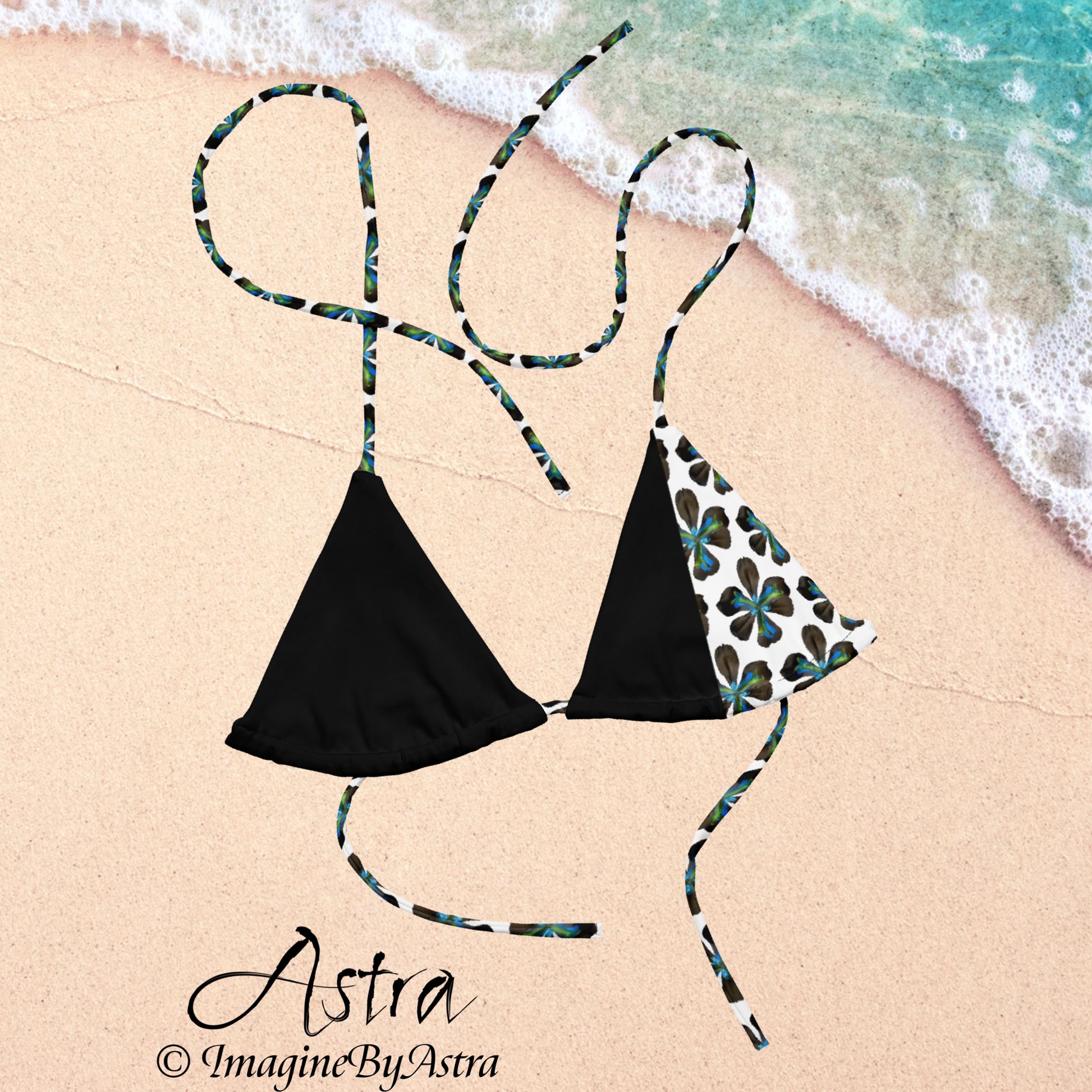 From the Astra Swimwear lineup: a black string bikini top with a white panel running down the side with a vibrant blue tropical flower print is laid out in the sand next to tropical waters, part of the Astra Swimwear Collection by designer, Imagine by Astra.