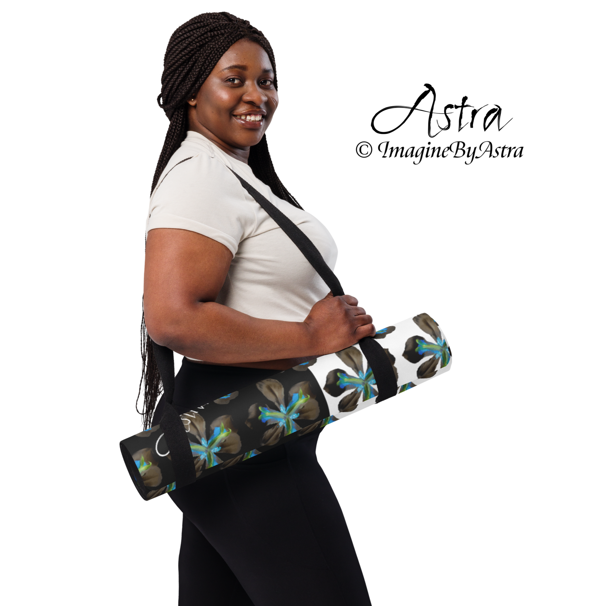 A black woman in a white t-shirt and black leggings carries a black and white yoga mat with a bright blue tropical flower print by the carrying strap included in the purchase, from Imagine by Astra Activewear.