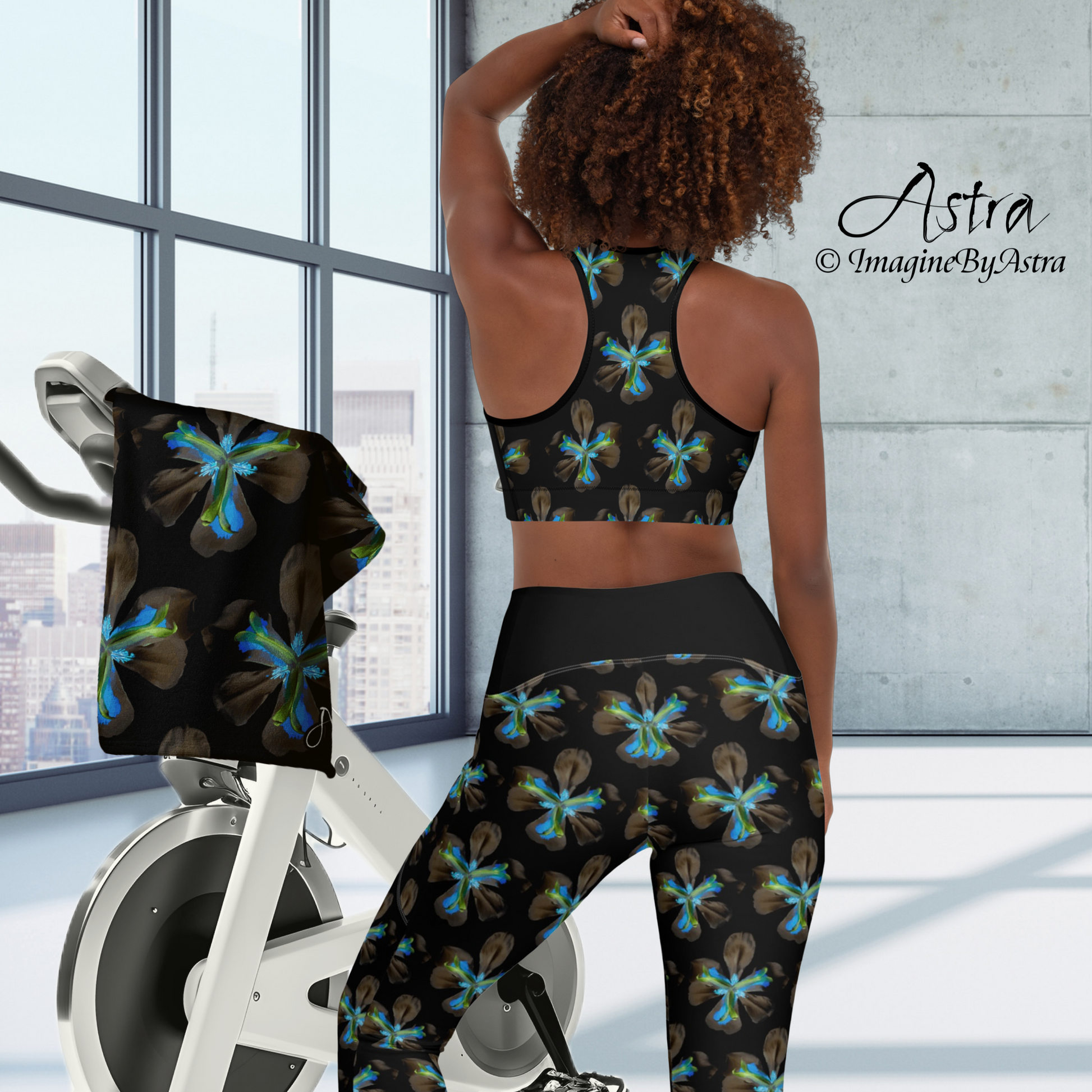 A fit medium brown skinned woman poses from behind wearing a black high waisted yoga legging and sports bra set with electric blue iris floral print from the Tropical Iris Collection by designer, ImagineByAstra. She stands before an exercise bike in a modern room with a large glass wall with city view. A matching black towel from the Tropical Iris Collection is draped over a handlebar of the bike.