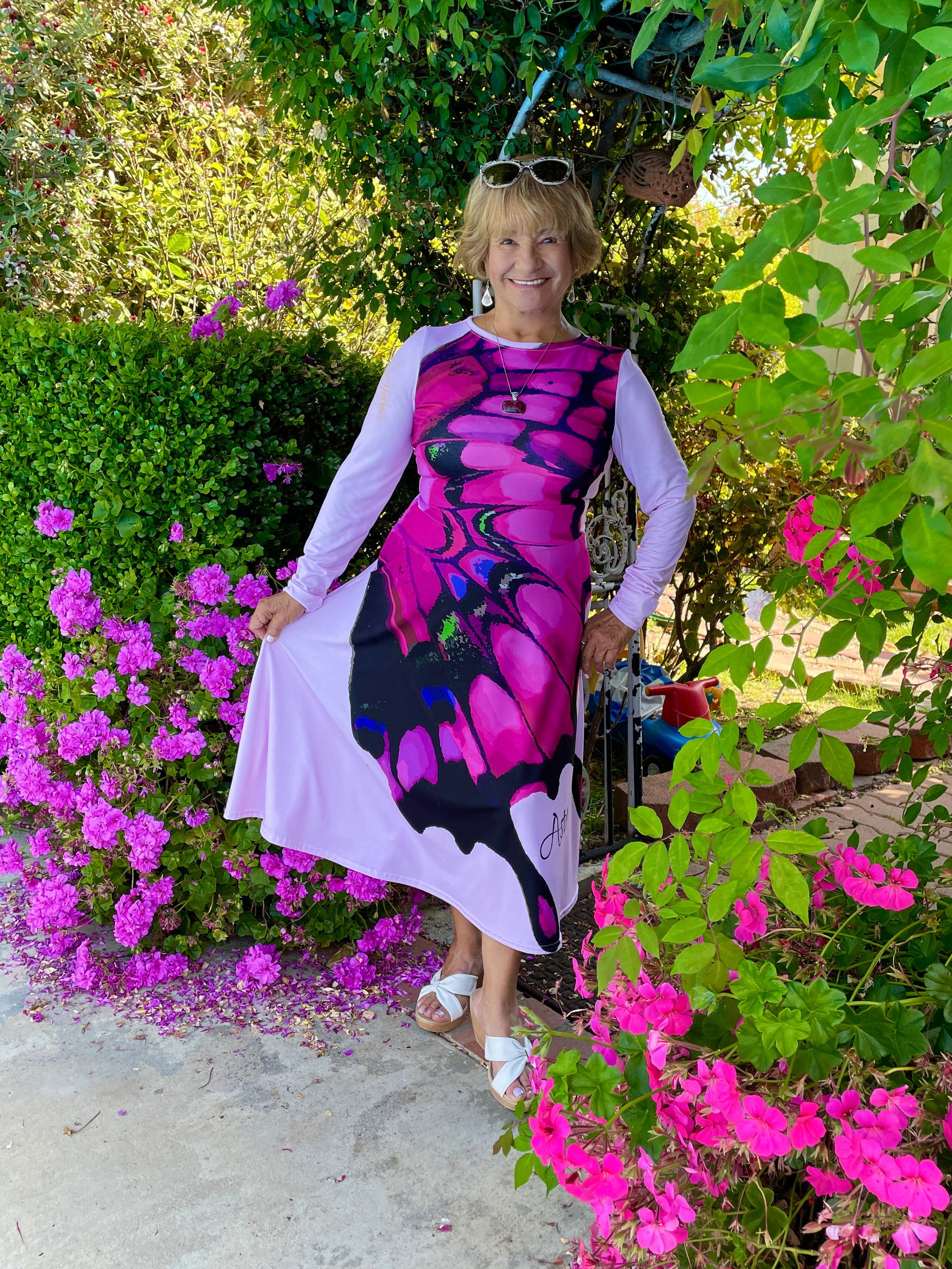 An olive skinned woman with short blond hair wears a long-sleeved, light pink, midi dress with a large pink butterfly graphic down the front by designer, Imagine by Astra. She stands in a garden archway surrounded by complementing pink flowers.