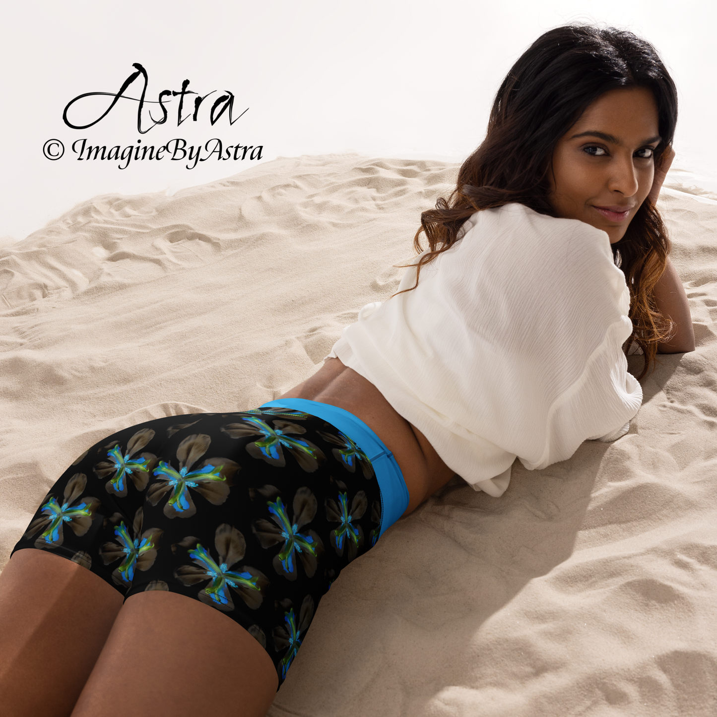 Back view of a brown skinned woman, lying on her stomach, wearing a pair of black high-waisted yoga shorts with a brilliant blue tropical flower print with matching blue waistband from the Tropical Iris Collection from designer, ImagineByAstra.