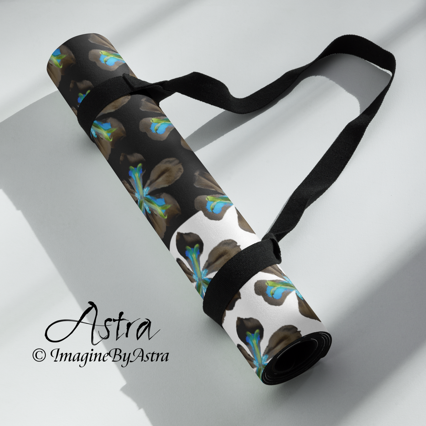 A yoga mat is rolled up with its accompanying carry strap. It bears a dramatic two toned black and white surface with a bright blue tropical flower print on top from the Tropical Iris Collection from Imagine by Astra Activewear.