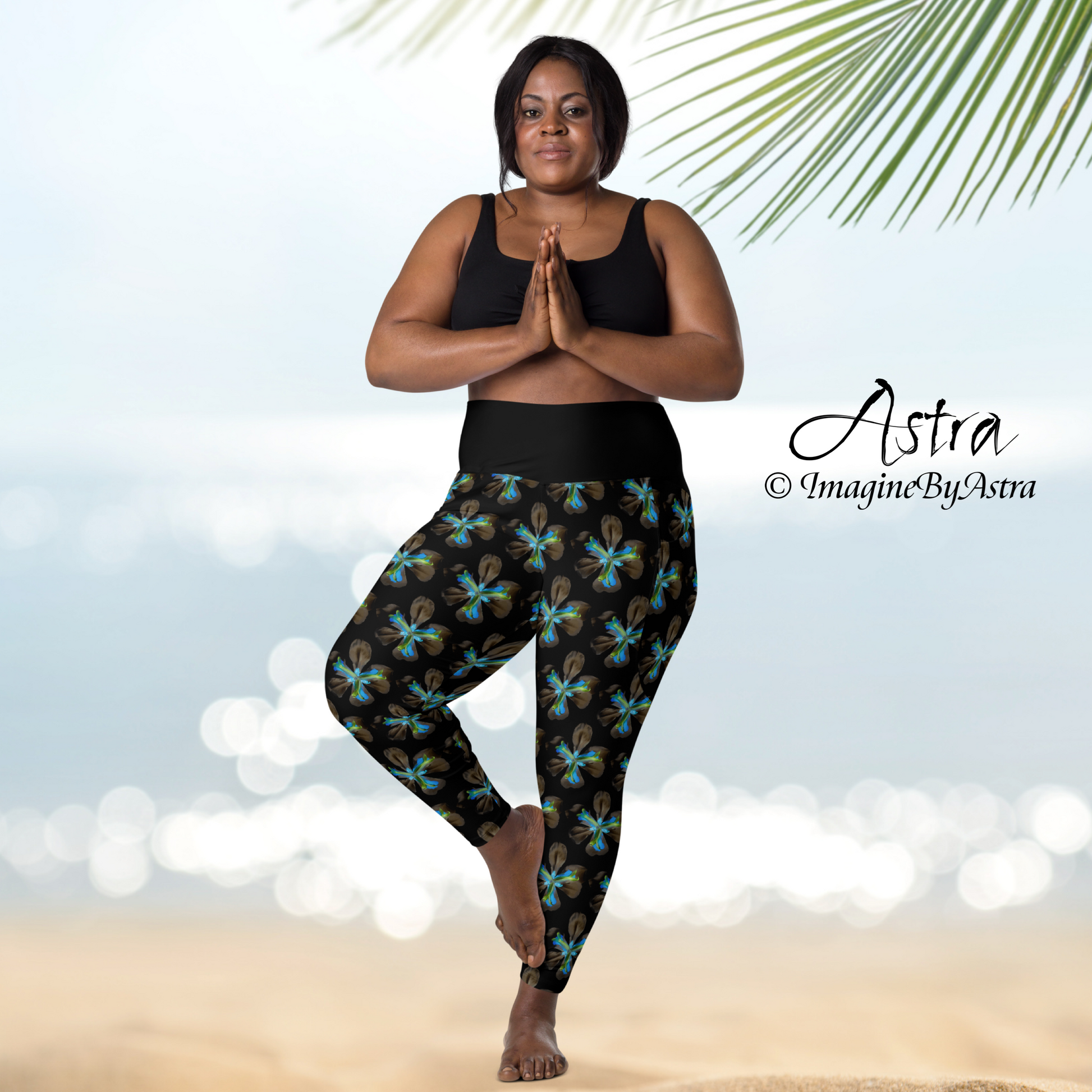 A young medium brown skinned, plus sized woman stands in prayer pose wearing high waisted black yoga leggings with a striking electric blue iris floral print and matching sports bra from the Tropical Iris Collection by designer, Imagine by Astra. A beach and ocean sparkle in the distance.