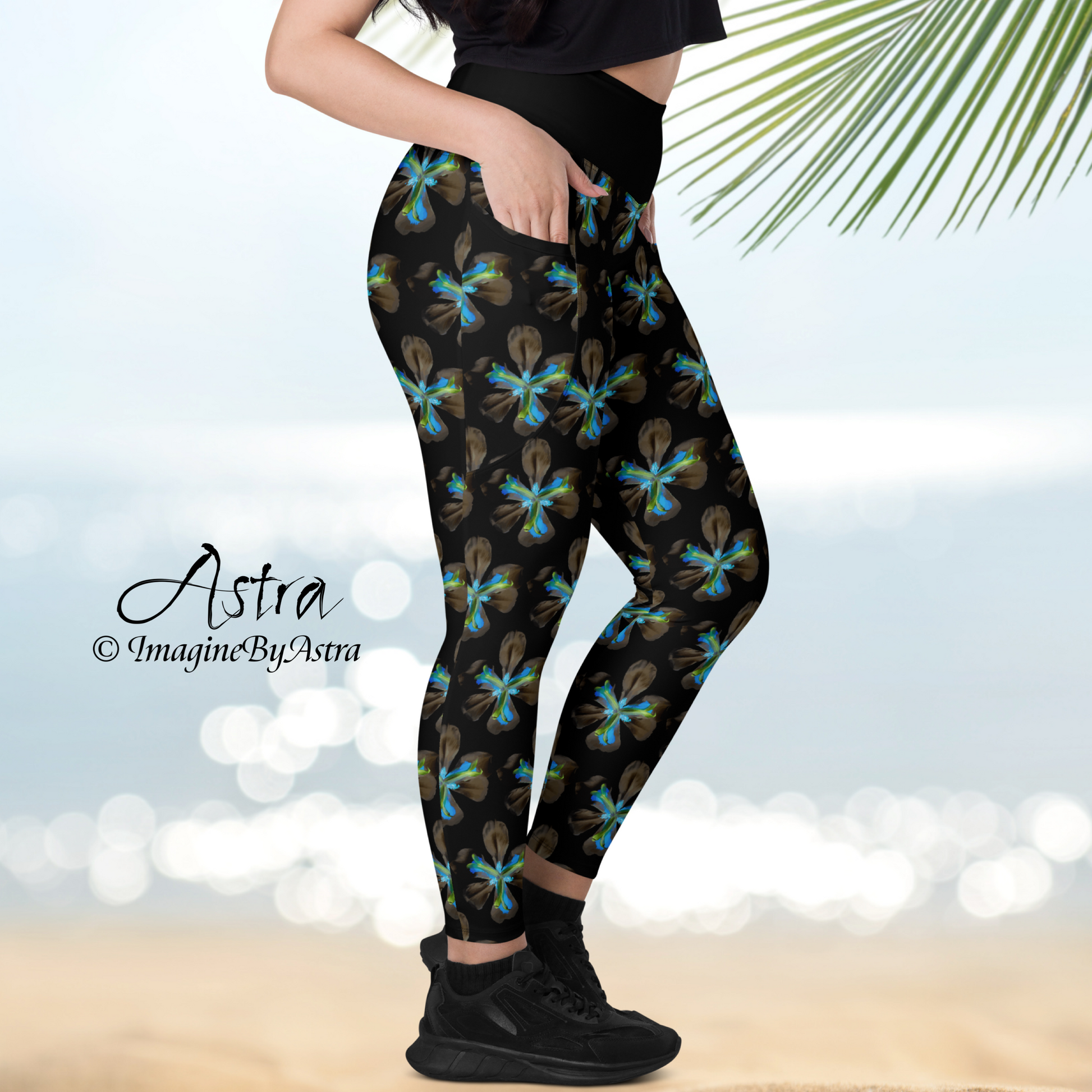 Right side view of the legs of a light skinned woman wearing black high waisted yoga leggings with a striking electric blue floral iris flower print from the Tropical Iris Collection by designer, Imagine by Astra. Her hand is in the pocket. The beach and ocean sparkle in the distance.