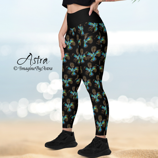 Left side view of the legs of a light skinned woman wearing black high waisted yoga leggings with a striking electric blue floral iris flower print from the Tropical Iris Collection by designer, Imagine by Astra. Her hand is on her hip, the beach and ocean sparkle in the distance.