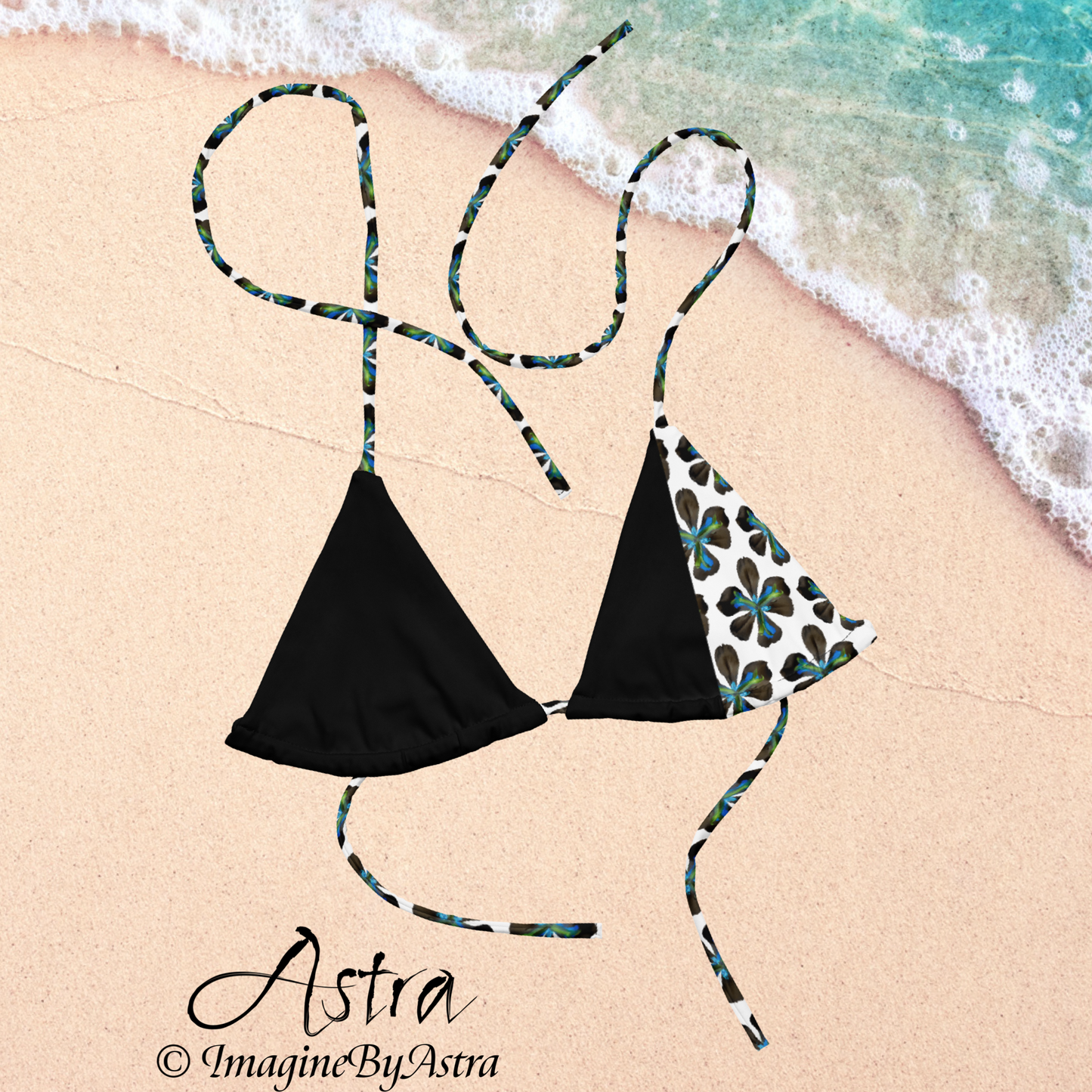 From the Astra Swimwear lineup: a black string bikini top with a white panel running down the side with a vibrant blue tropical flower print is laid out in the sand next to tropical waters, part of the Astra Swimwear Collection by designer, Imagine by Astra. 