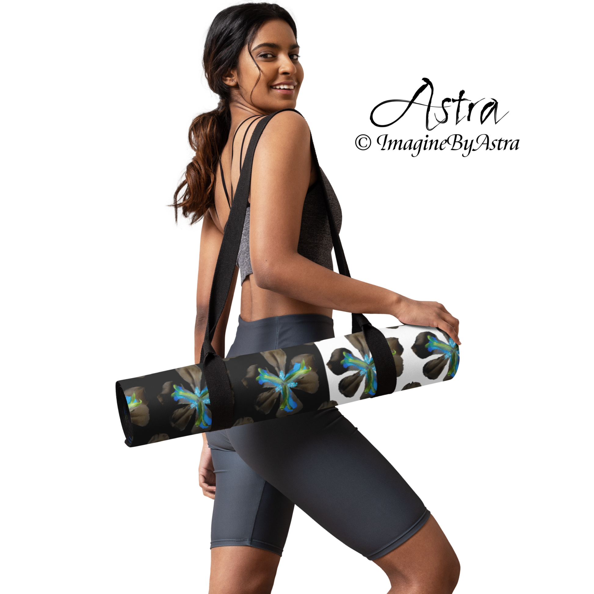 A black woman carries a black and white yoga mat with a bright blue tropical flower print with a carrying strap included in the purchase, from activewear brand, Astra activewear.