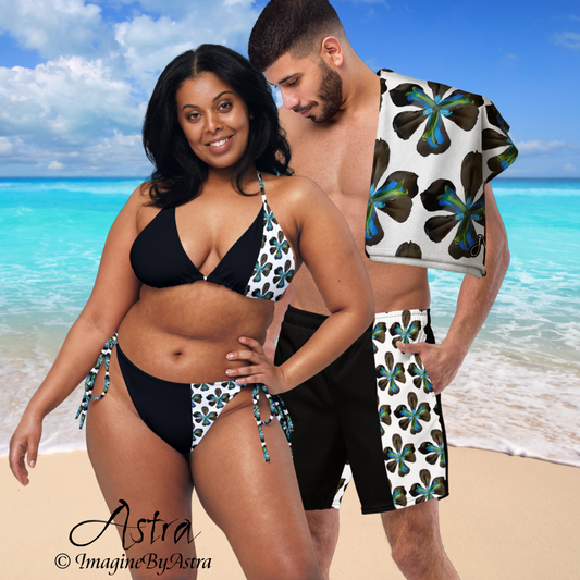 A black man and woman couple wear matching black and white swimsuits with tropical flower print on a tropical beach: she wears a black string bikini with a white offset panel with tropical flower print. He wears black swim trunks with a white offset panel with tropical flowers. Part of the Tropical Iris Swimwear Collection by designer Imagine by Astra.