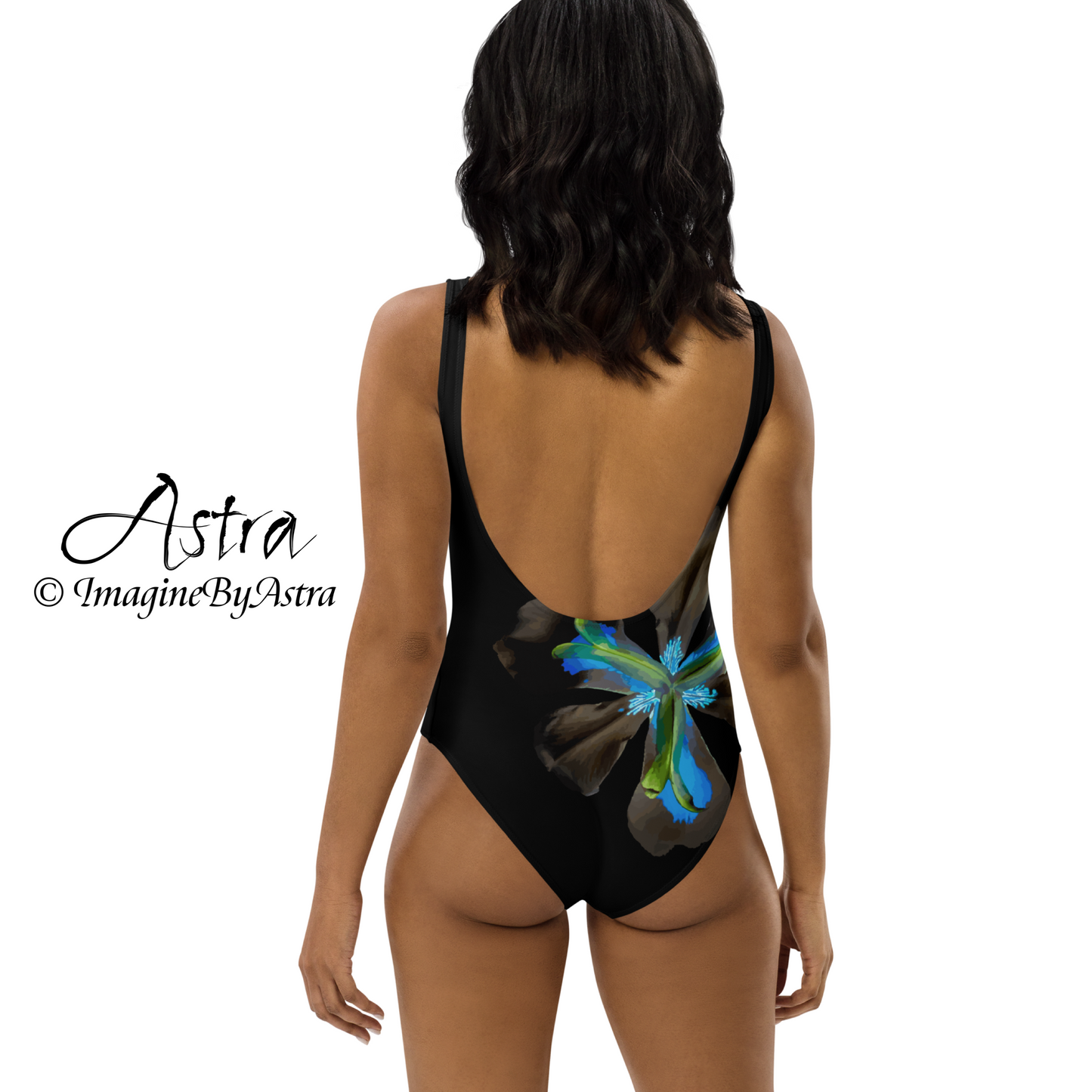 From the Astra Swimwear lineup: the back of a black woman is seen wearing a black one-piece swimsuit with a large vibrant blue tropical flower graphic down one side. The swimsuit has a flattering backless cut and high cut at the hips. Part of the Tropical Iris Collection by designer, Imagine By Astra.