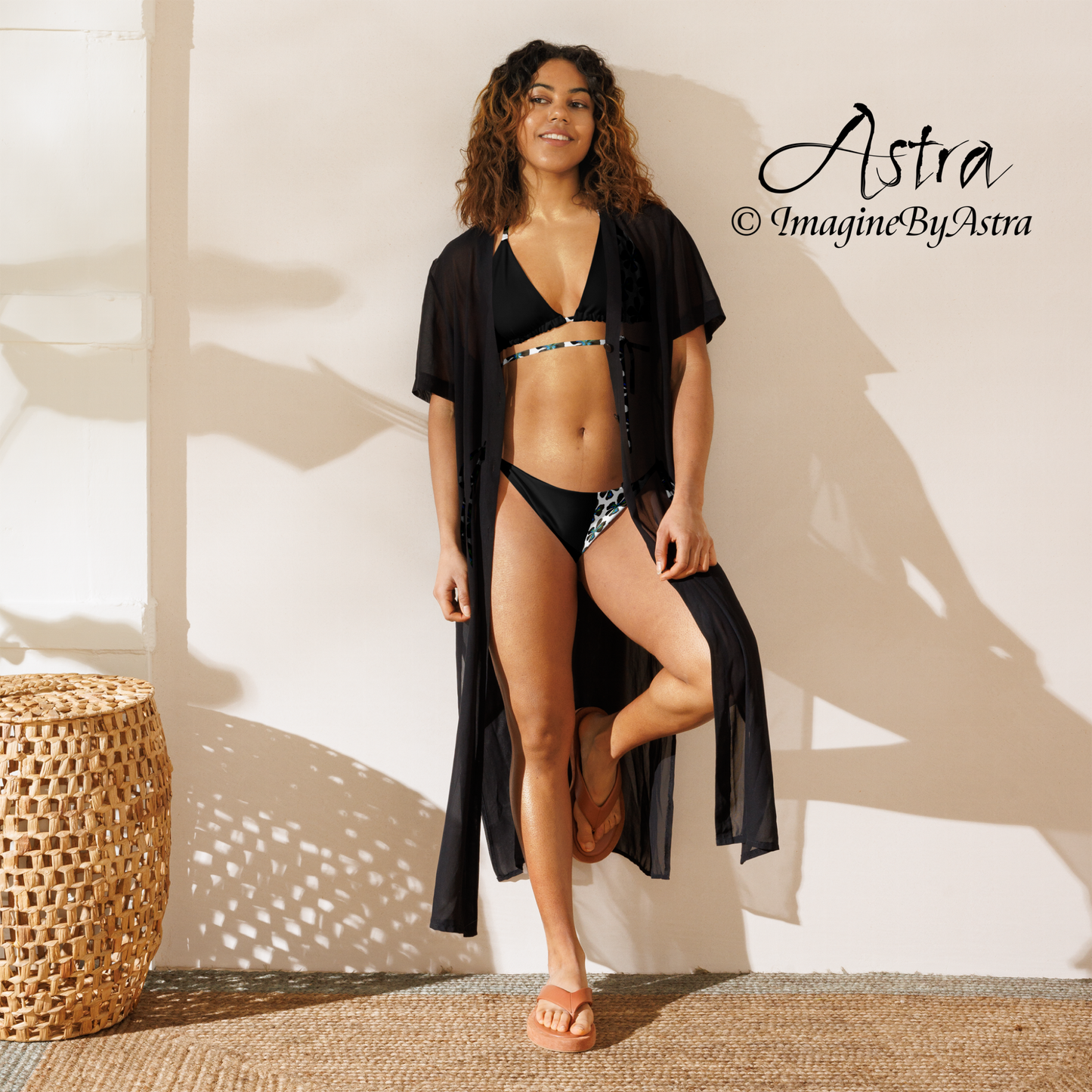 A slender black woman leans up against a wall, wearing a black and white string bikini with a blue tropical flower print with the string creatively wrapped around the waist, part of the Tropical Iris Collection by designer, ImagineByAstra. From the Astra Swimwear lineup.