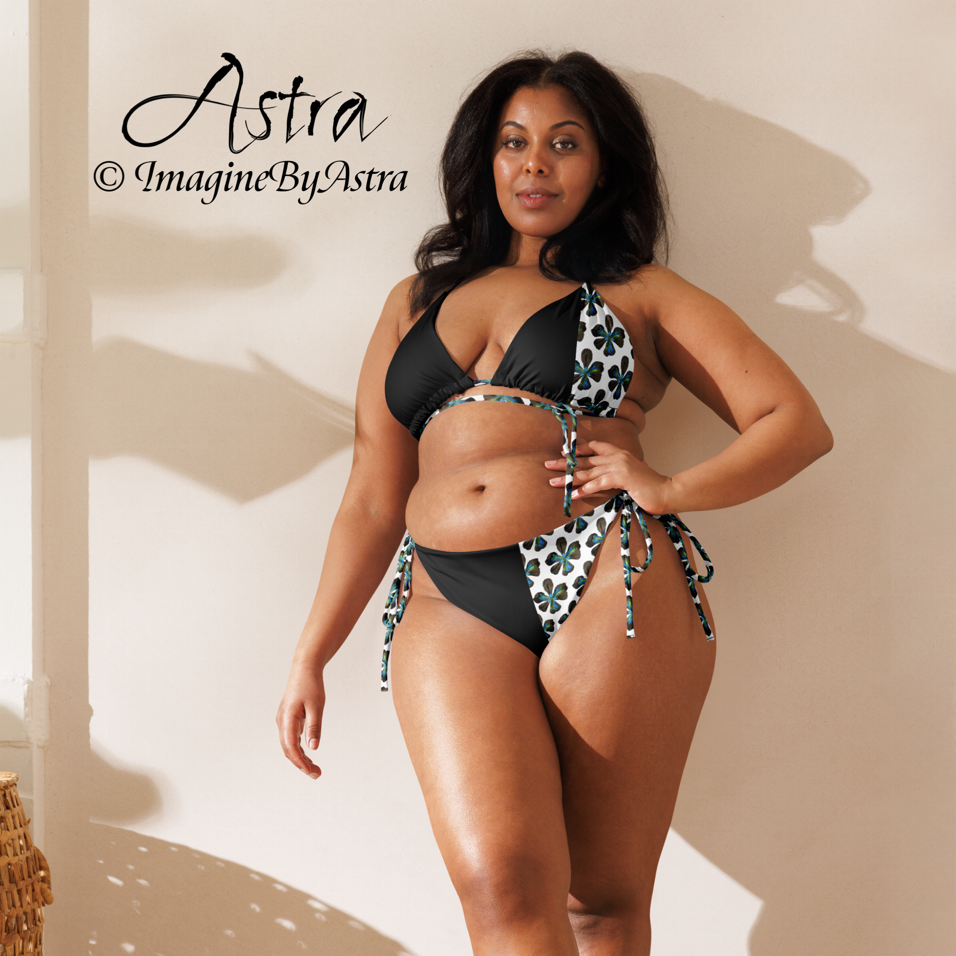 A plus size black woman leans up against a wall, wearing a black and white string bikini with a blue tropical flower print with the string creatively wrapped around the waist, part of the Tropical Iris Collection by designer, ImagineByAstra. From the Astra Swimwear lineup.