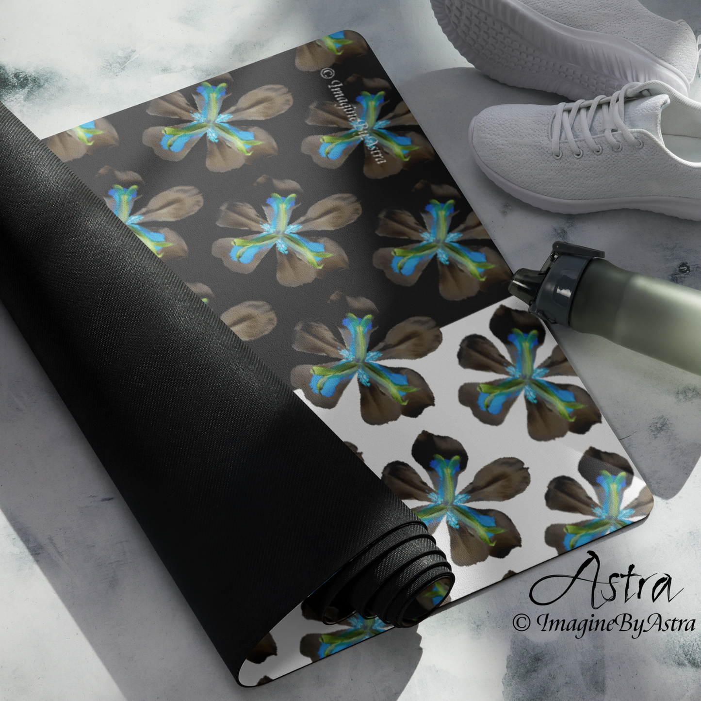 A yoga mat is partially unrolled revealing a dramatic two toned black and white surface with a bright blue tropical flower print on top, the black non slip rubber back is shown on the back of the mat. From brand, Imagine by Astra Activewear.