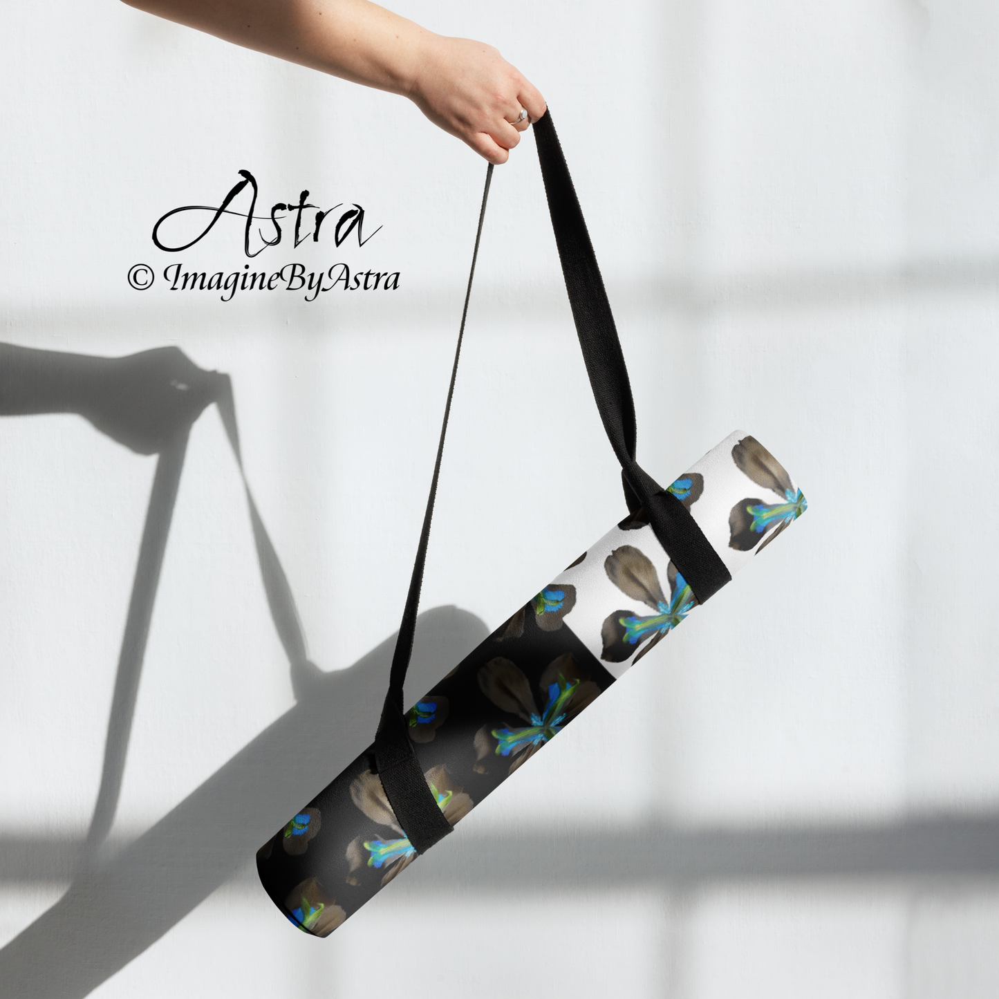 A yoga mat is rolled up and dangled by its accompanying carry strap. It bears a dramatic two toned black and white surface with a bright blue tropical flower print on top from the Tropical Iris Collection from Imagine by Astra Activewear.