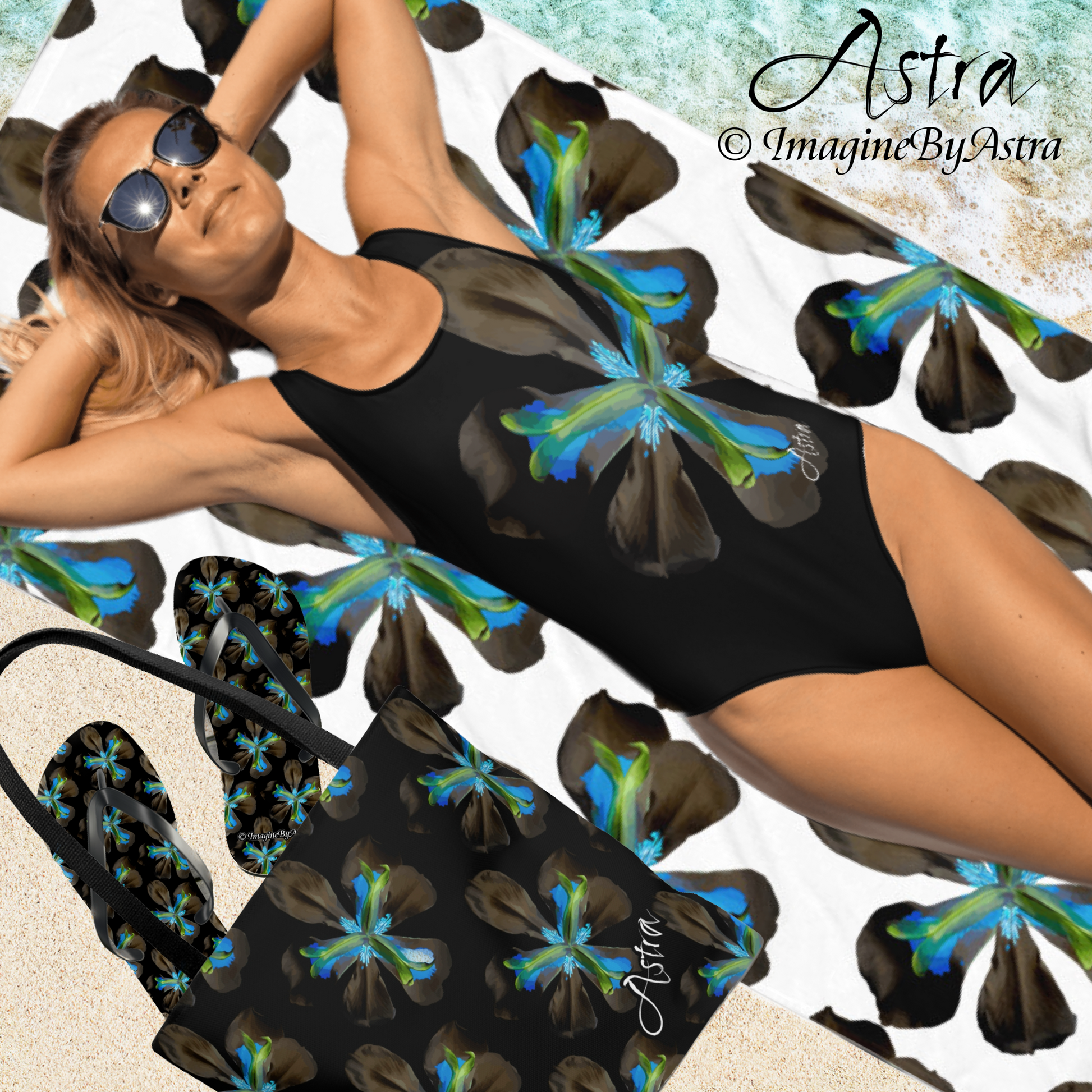 A white blond haired woman lays on her back on a beach, wearing a black one-piece swimsuit with a large vibrant blue tropical flower graphic down the front from the Astra Swimwear collection. She lies on a matching towel and is flanked by matching tote bag and flip flops from the Tropical Iris Collection by designer, Imagine By Astra.